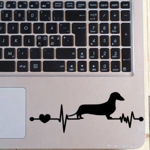 May include: A black silhouette of a dachshund with a heart and a heartbeat line on a silver laptop.