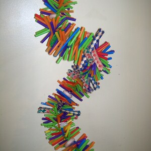 May include: A colorful, multi-layered bird toy made from plastic drinking straws. The toy is strung together with beads and has a red plastic flower at the bottom.