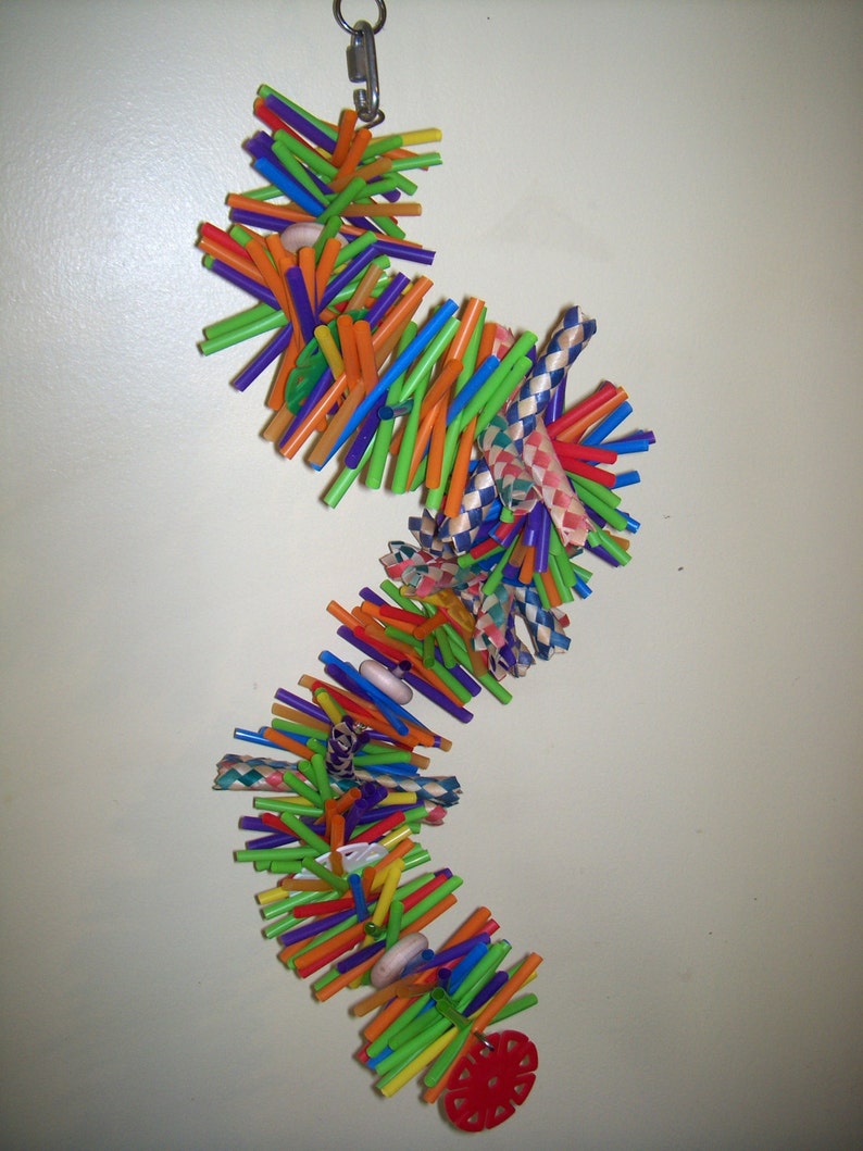 May include: A colorful, multi-layered bird toy made from plastic drinking straws. The toy is strung together with beads and has a red plastic flower at the bottom.