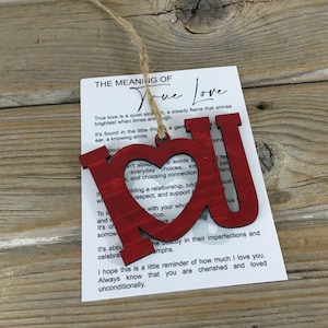 May include: Red wooden cutout of the letters "I" and "U" with a heart in the middle. The letters are attached to a card with the text "The Meaning of True Love" and a description of what true love means.