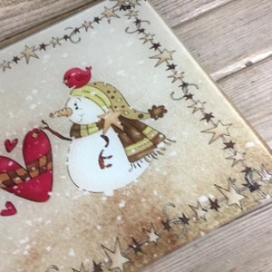 K&ouml;nnte beinhalten: Ein Schneemann mit einem roten Vogel auf dem Kopf h&auml;lt ein rotes Herz mit einem karierten Muster. Der Hintergrund ist cremefarben mit einem braunen Sternrand.