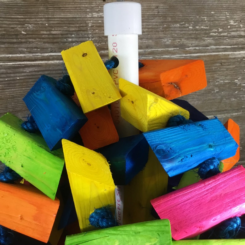 May include: A colorful wooden bird toy with a variety of shapes and sizes. The toy is made of brightly colored wood and is strung together with rope. The toy is attached to a white PVC pipe.