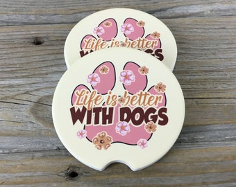 Puede incluir: Dos posavasos redondos de color crema con un diseño de huella de pata rosa. Los posavasos tienen un texto marrón que dice "Life is better with dogs" y están decorados con flores rosas y marrones.