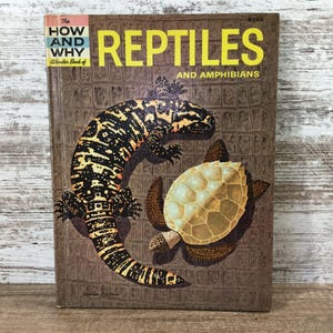 May include: A hardcover book titled The How and Why Wonder Book of Reptiles and Amphibians. The brown cover features a large black and yellow Gila monster and a light-colored turtle illustration against a textured, grid-like background pattern.