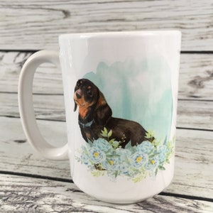 May include: White ceramic coffee mug with a brown and black dachshund dog illustration. The dog is surrounded by blue watercolor flowers.