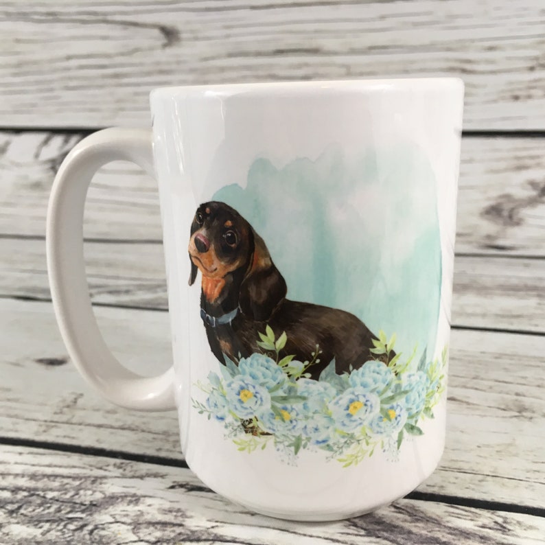May include: White ceramic coffee mug with a brown and black dachshund dog illustration. The dog is surrounded by blue watercolor flowers.