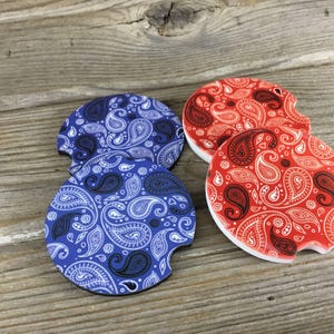 May include: Four car coasters with a paisley design. Two are blue with white and black paisley patterns, and two are red with white and black paisley patterns. The coasters are on a wooden surface.