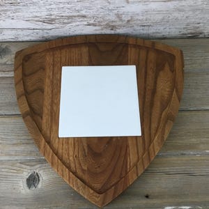 May include: A wooden shield-shaped serving board with a recessed center. A square, white tile is set in the center of the board. The wood has a warm, brown tone, and the board has a carved edge.