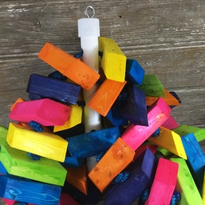 May include: A colorful wooden toy for birds, featuring a variety of brightly colored blocks attached to a white pole. The blocks are in shades of blue, green, yellow, orange, pink, and red.