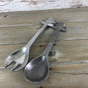 May include: Two silver-colored salad servers. One server is a spoon, and the other is a fork. Both have a tree-shaped design at the end of the handle. The servers are made of metal and rest on a wooden surface.