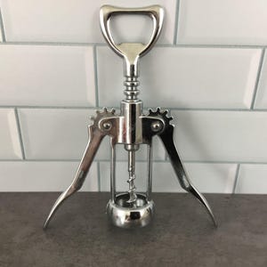 May include: A polished, silver-toned wine bottle opener featuring a handle, corkscrew, and levers. The opener has a gear-like design on the sides and is designed to extract corks from wine bottles. The tool is made of metal.