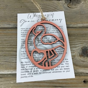 May include: A pink wooden flamingo ornament with a heart cutout. The ornament is hanging from a twine string. The text on the card reads "Whispers of Grace & Harmony".