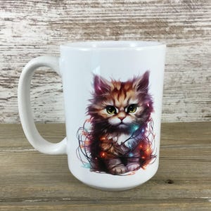 May include: White ceramic mug featuring a fluffy brown and tan kitten with green eyes, tangled in colorful Christmas lights. The mug has a large handle and is suitable for hot beverages.