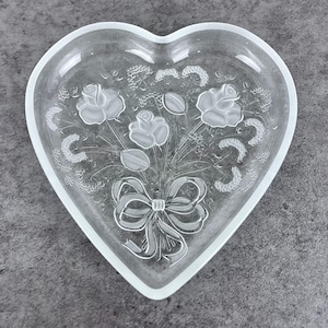 May include: Clear glass heart-shaped dish with a frosted edge. The dish features an etched design of a bouquet of roses tied with a bow. The roses, leaves, and bow are all etched into the glass, creating a delicate, detailed pattern.