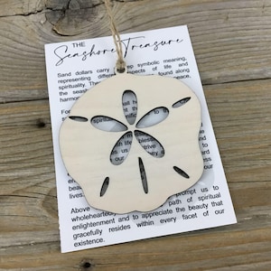 May include: A wooden sand dollar ornament with a cutout design. The ornament is attached to a string and has a card with the text "Seashore Treasure" and a description of the symbolic meaning of sand dollars.