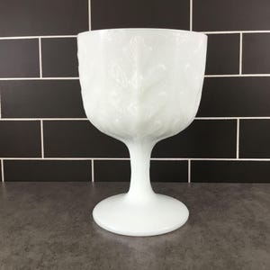May include: A white, stemmed goblet with a wide bowl and a round base. The bowl features a detailed leaf pattern. The goblet is made of opaque glass and stands on a dark gray surface against a black tiled background.