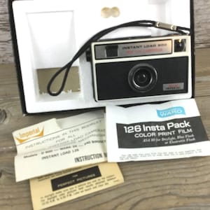 May include: A vintage Imperial Instant Load 126 camera in its original box, with a black and white design. The camera has a lens, flash, and a black strap. Included are instruction booklets and a film pack.