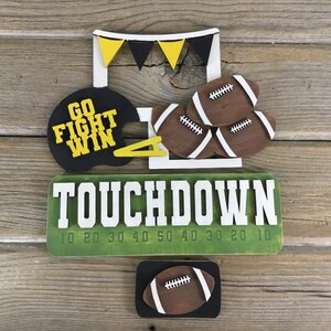 Peut inclure: Panneau en bois avec les mots "TOUCHDOWN" en lettres blanches sur fond vert. Le panneau comporte également un terrain de football avec des lignes de yardage marquées de 10, 20, 30, 40, 50, 40, 30, 20, 10. Il y a un casque de football avec les mots "GO FIGHT WIN" dessus et un poteau de but de football avec trois ballons de football dans la zone d'en-but.