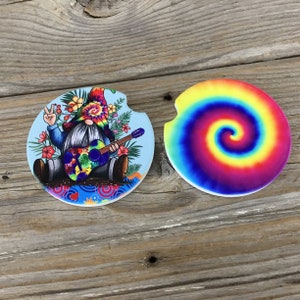 May include: Two round ceramic coasters with a tie-dye design. One coaster features a gnome playing a guitar with a peace sign, surrounded by flowers. The other coaster has a colorful spiral tie-dye pattern.