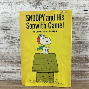 May include: A vintage book titled "SNOOPY and His Sopwith Camel" by Charles M. Schulz. The cover is yellow and features Snoopy in a pilot's cap and scarf, sitting atop his doghouse. The book's cover is slightly worn.