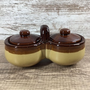 May include: A double-chambered ceramic condiment server featuring a two-tone glaze with a dark brown upper section and a light tan lower section. The piece includes two matching round lids with small knobs and a central vertical handle for carrying.