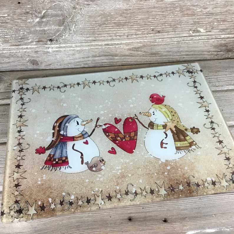 K&ouml;nnte beinhalten: Ein Schneidebrett aus Glas mit einer Winterszene, die zwei Schneem&auml;nner zeigt, die ein rotes Herz halten. Die Schneem&auml;nner tragen Schals und H&uuml;te. Der Hintergrund ist beigefarben mit einem Rand aus braunen Sternen.