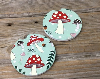 May include: Two round, light blue coasters with a white background and red and white mushroom illustrations. The mushrooms have white stems and red caps with white spots. The coasters have a green leafy pattern around the mushrooms.