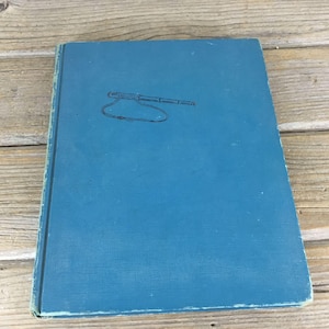 May include: A vintage book with a teal cover. The cover features a simple line drawing of a telescope with a rope. The book's edges show signs of wear, suggesting its age. The background is a wooden surface.