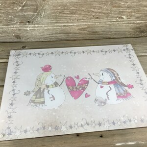 K&ouml;nnte beinhalten: Ein Schneidebrett aus Glas mit einer Winterszene, die zwei Schneem&auml;nner zeigt, die ein rotes Herz halten. Die Schneem&auml;nner tragen Schals und H&uuml;te. Der Hintergrund ist hellbraun mit einem wei&szlig;en Sternrand.