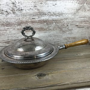 May include: A silver-colored serving dish with a lid and a wooden handle. The dish has a decorative handle on the lid and a detailed rim. The wooden handle is attached to the side of the dish.