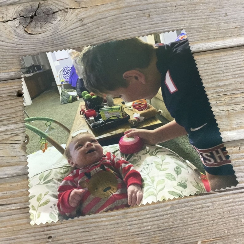 May include: A smiling baby in a red and white striped onesie with a brown bear design is being looked at by an older sibling wearing a black and red shirt with the word "SH" on it. The baby is sitting on a white and green patterned pillow.