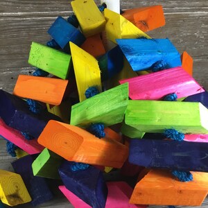 May include: A colorful wooden toy for birds, made of brightly colored blocks in shades of blue, green, orange, pink, and yellow. The blocks are connected together with string.