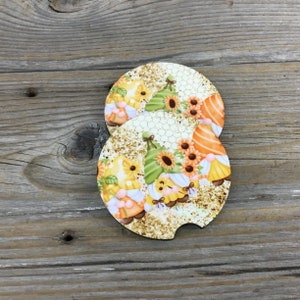 May include: Two round, white, cork coasters with a honeycomb pattern and a design of gnomes wearing hats and holding sunflowers. The gnomes are surrounded by glitter.