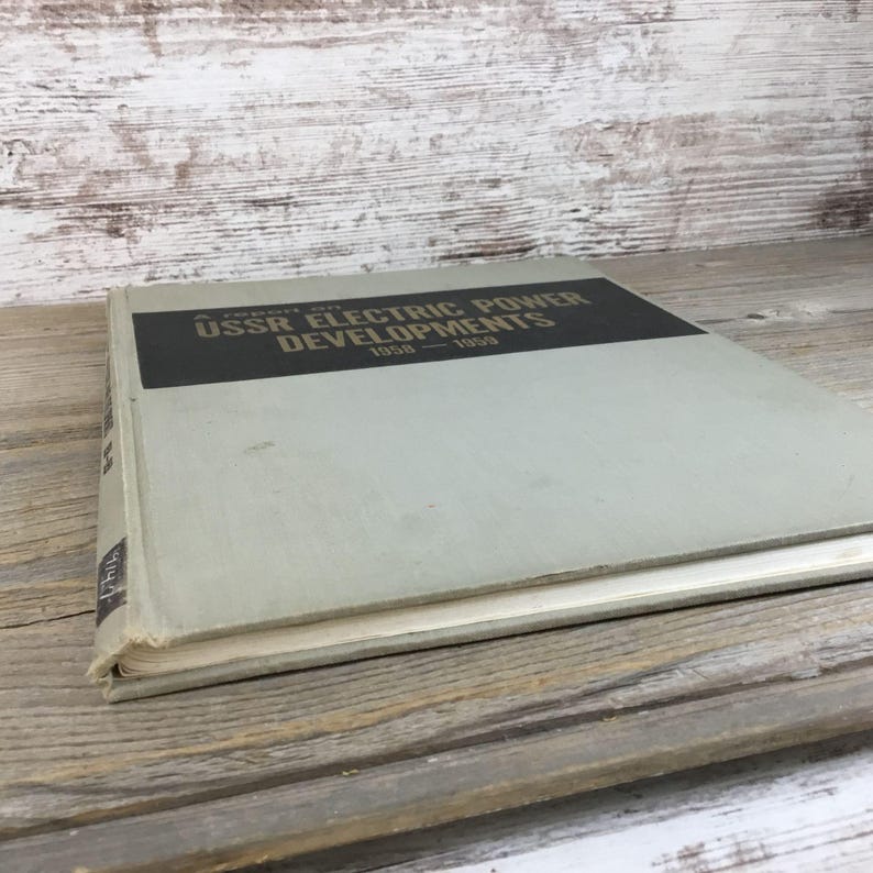 以下が含まれることがあります： 「A Report on USSR Electric Power Developments 1958-1959」と題されたヴィンテージ本。本は、黒いテキストが入った薄い灰色の表紙です。本は、風化した木製の表面に置かれています。