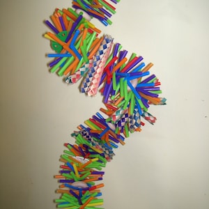 May include: A colorful, multi-layered toy made of woven straw, plastic drinking straws, and plastic buttons. The toy is shaped like a foot and is hanging from a silver hook.