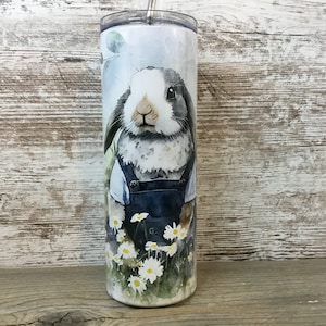 May include: A stainless steel tumbler with a white background and a watercolor illustration of a rabbit wearing overalls and standing in a field of daisies.