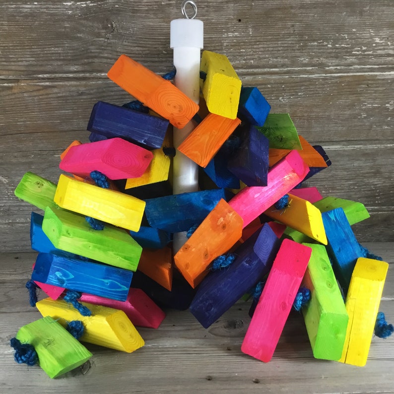 May include: A colorful wooden toy for birds, featuring a variety of brightly colored blocks connected by rope and a white wooden dowel. The blocks are painted in shades of blue, green, yellow, orange, pink, and lime green.