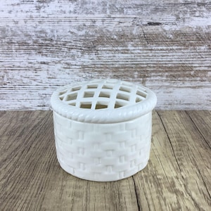 May include: A white ceramic flower frog with a basket weave design. The lid has a grid pattern for arranging flowers. The rim of the lid has a rope-like texture. The container sits on a wooden surface.