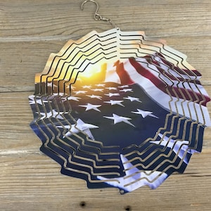 May include: A metal wind spinner featuring a design of an American flag waving in the wind. The flag is red, white, and blue with white stars on a blue field. The background is a sunset.