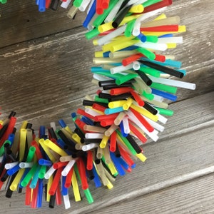 Pu&ograve; includere: Una ghirlanda colorata realizzata con cannucce da bere di vari colori, tra cui rosso, giallo, blu, verde e nero. Le cannucce sono disposte in un motivo circolare, creando una decorazione festosa e fantasiosa.