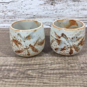 May include: Two small, cylindrical ceramic cups featuring a speckled off-white glaze with hand-painted brown, leaf-like patterns. The rims are accented with a thin, warm brown wash, and the overall finish has a textured, earthy appearance.