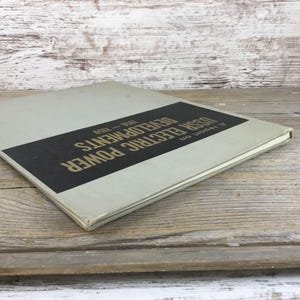 以下が含まれることがあります： 「USSR Electric Power Developments 1958-1959」というタイトルのヴィンテージ本。薄いグレーの表紙と黒い長方形のラベルが付いています。テキストは金色です。本は風化した木製の表面に置かれています。