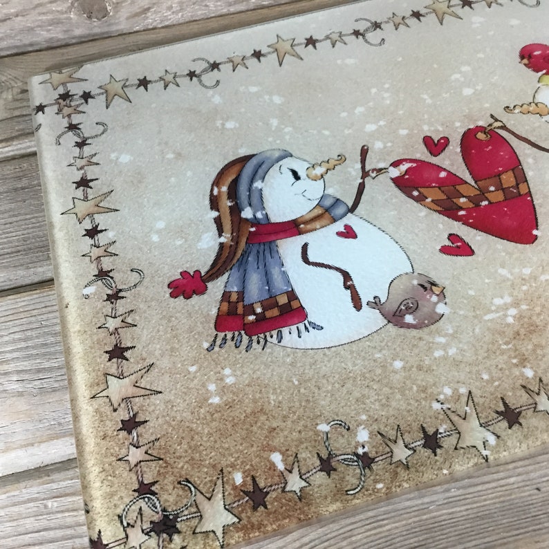 K&ouml;nnte beinhalten: Ein Schneemann mit einem blauen Schal und roten F&auml;ustlingen h&auml;lt ein rotes Herz mit einem braunen und wei&szlig;en Karomuster. Ein kleiner brauner Vogel sitzt auf dem Arm des Schneemanns. Der Hintergrund ist beige mit einem Rand aus braunen Sternen.