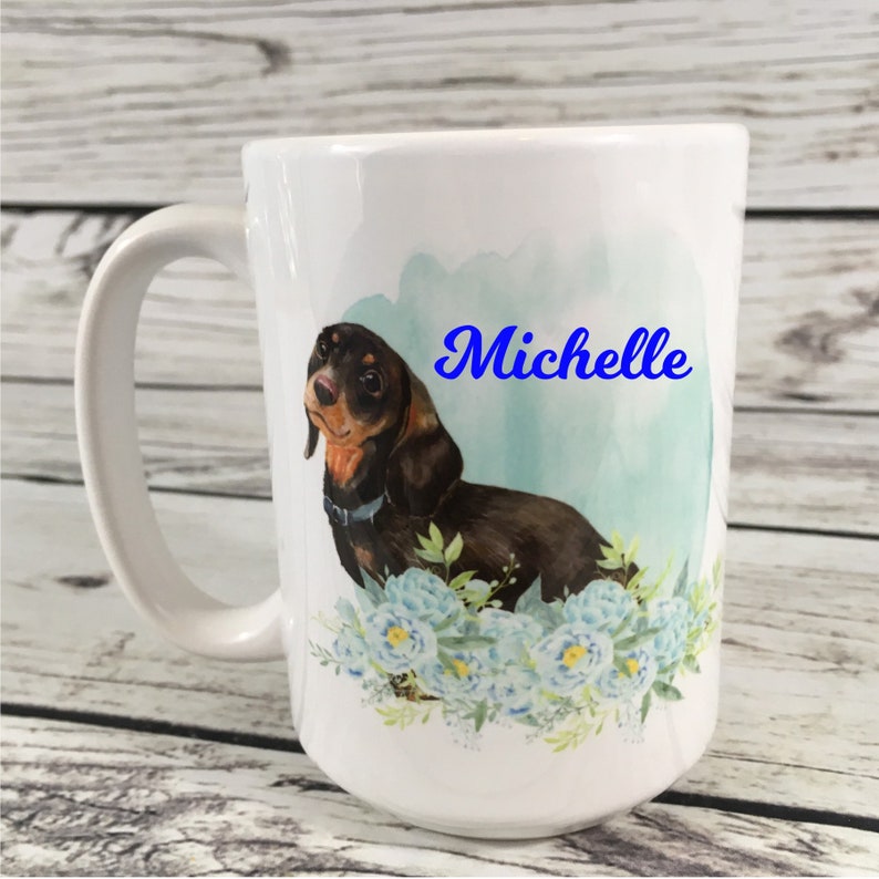 May include: White ceramic mug with a watercolor design of a dachshund dog with blue flowers. The mug has the name "Michelle" written in blue cursive.