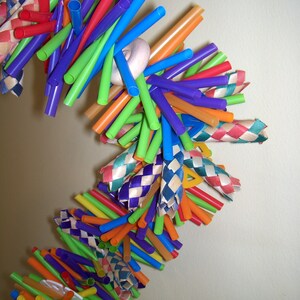 May include: A colorful, woven, hanging toy made from drinking straws and ribbon. The toy is made of various colors, including red, orange, yellow, green, blue, purple, and white. The toy is hanging from the ceiling and is swaying gently in the breeze.