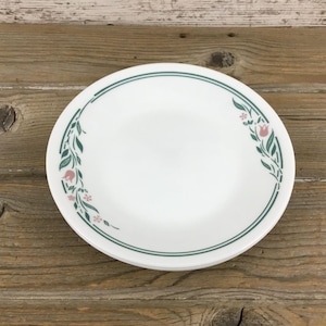 May include: White ceramic plate with a green and pink floral design around the rim. The plate has a double green line border. The floral pattern includes green leaves and pink tulip-like flowers. The plate is on a wooden surface.