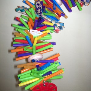 May include: A colorful toy made of plastic drinking straws, with a red plastic ring and a wooden wheel. The toy is designed for birds to play with.