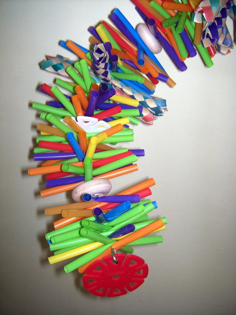 May include: A colorful toy made of plastic drinking straws, with a red plastic ring and a wooden wheel. The toy is designed for birds to play with.