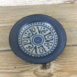 May include: A small, round, blue ceramic plate with the word "love" repeated in a circle in a lighter blue color. The plate has a raised edge and a simple, rustic design.