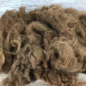 May include: A pile of brown alpaca fiber, with a soft, fluffy texture. The fibers are a warm, earthy brown color, and appear to be unspun. The background is a light-colored, weathered wood surface.